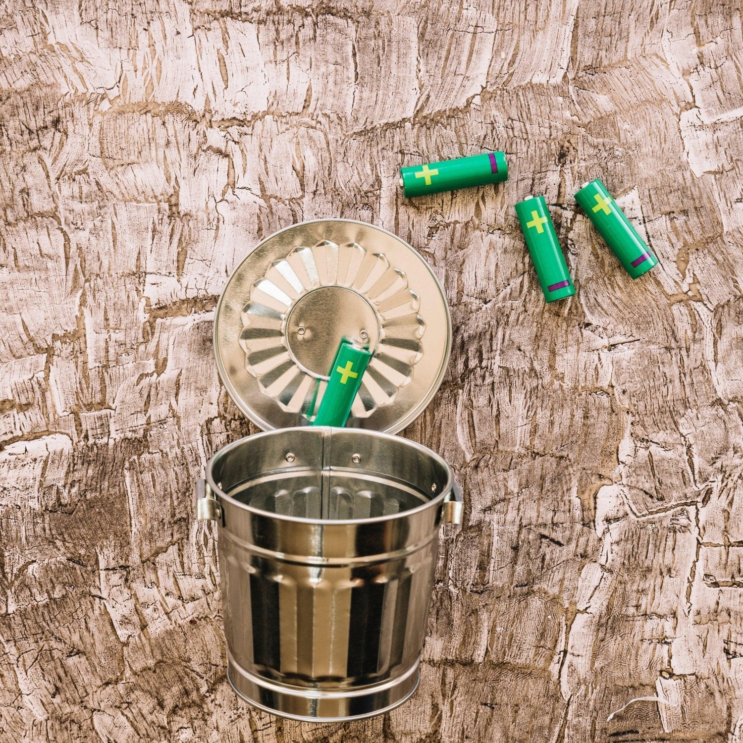 Green batteries discarded in metal trash can on textured wood surface, highlighting battery disposal and recycling